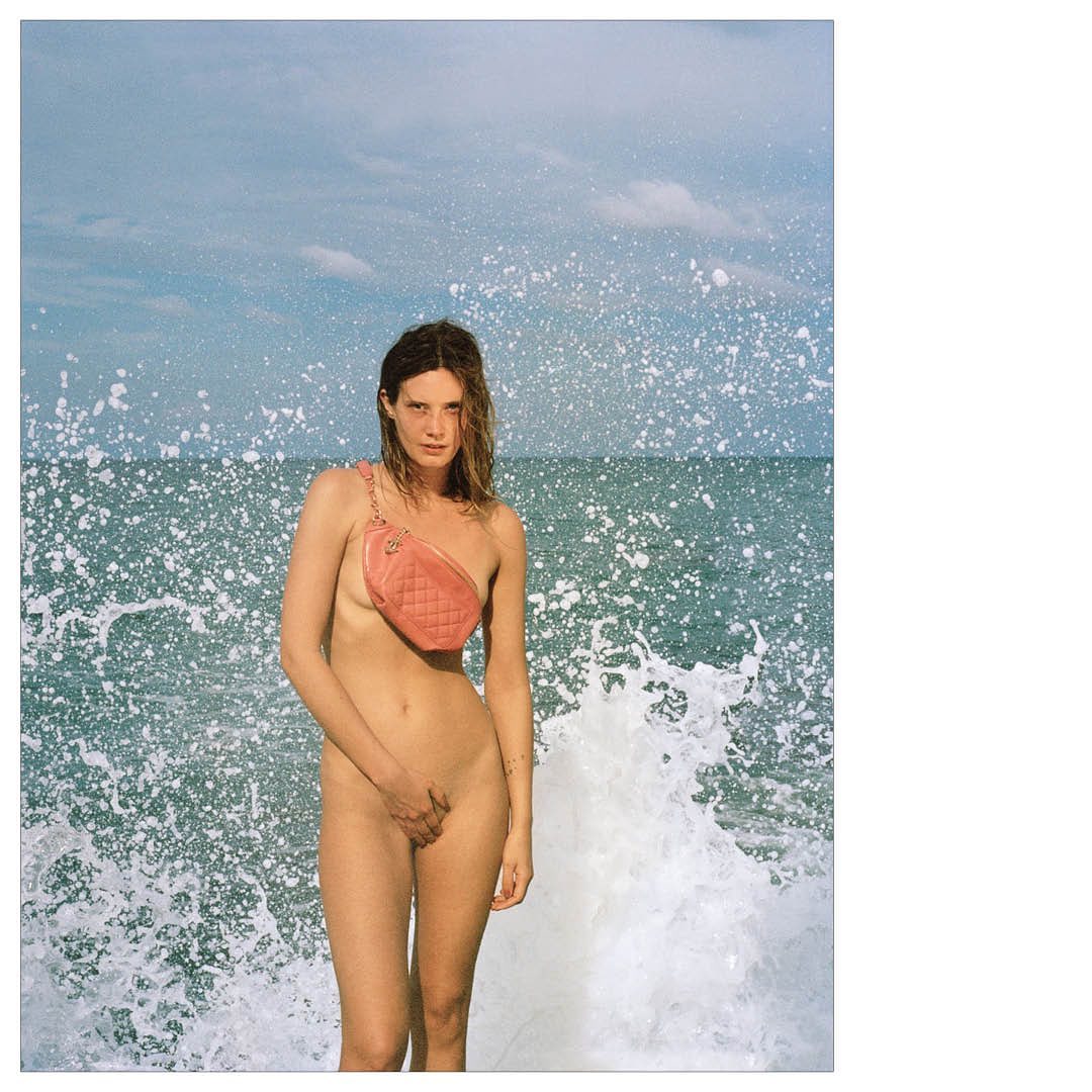 Nude woman on a beach stands in front of crashing wave wearing a pink quilted fanny pack with a gold chain over her shoulder.