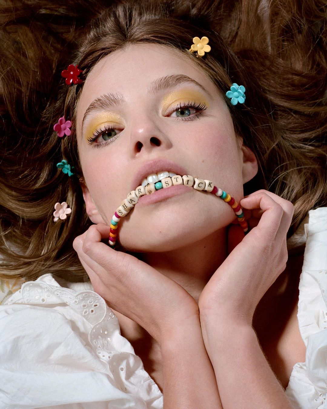 Close-up portrait of a young woman with floral hair clips, holding a beaded necklace in her mouth, and wearing yellow eyeshadow.
