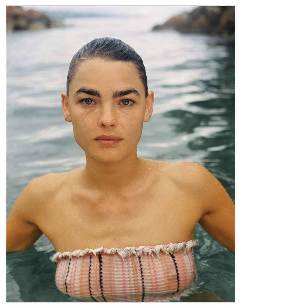 Portrait of a young woman with short hair in a patterned tube top, partially submerged in rippling water, with a distant shoreline.