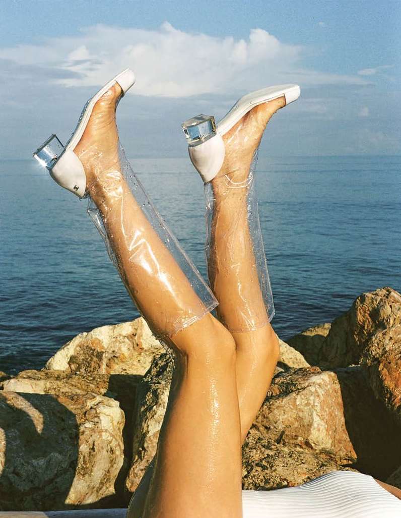Woman's legs wearing clear, high-heeled shoes against a backdrop of the sea and sky, styled with a beach setting and clear blue water.
