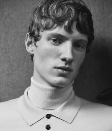 Close-up portrait of a young man with wavy brown hair wearing a turtleneck and buttoned polo shirt in black and white.
