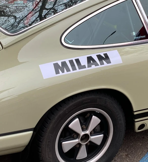 Close-up of a beige Porsche with a black stripe and a 'Milan' decal above a black tire with a silver hubcap, providing an automotive detail.