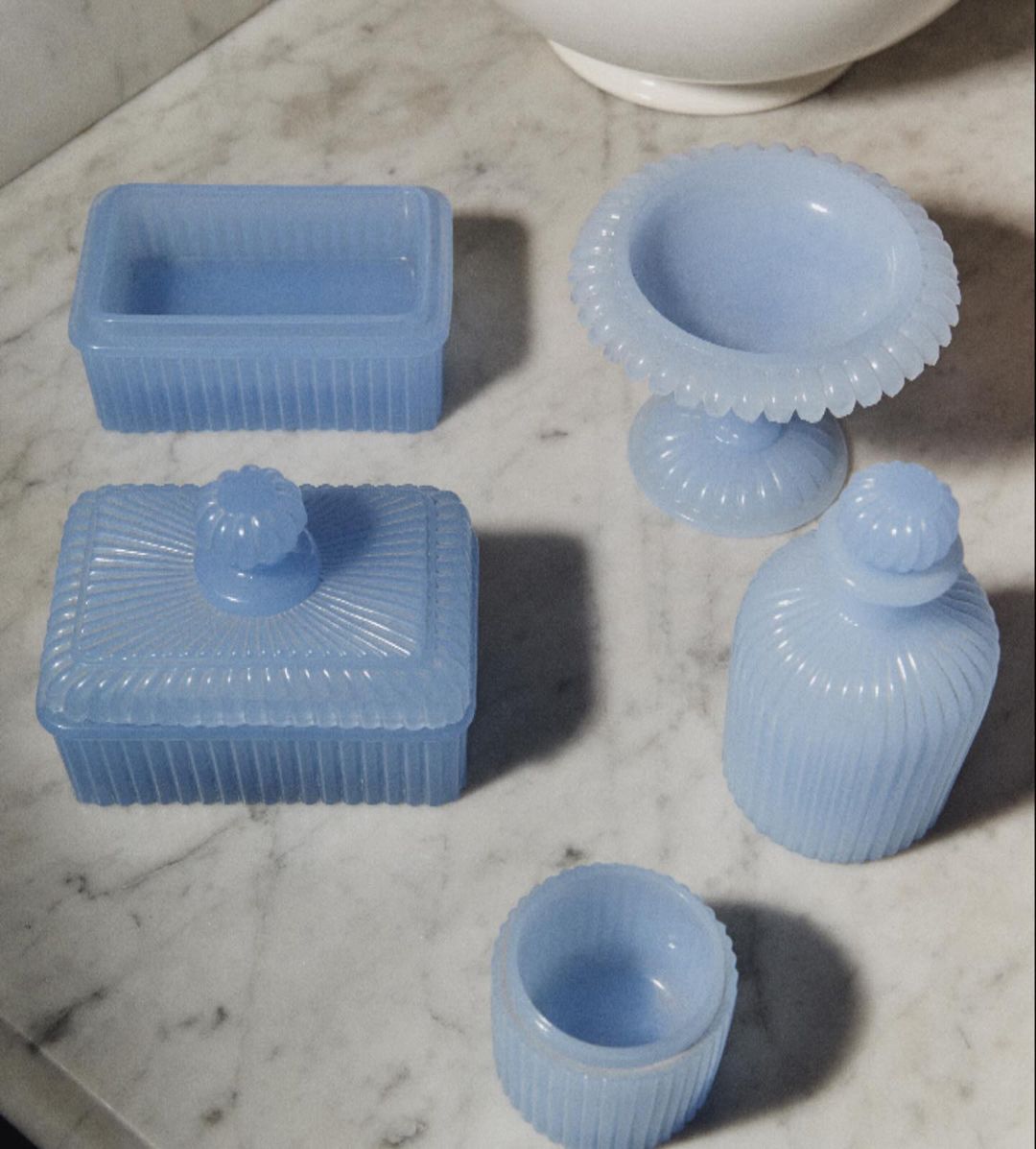 Collection of light blue vintage-style vanity objects, including covered boxes, a pedestal bowl, bottle, and cup, on a marble surface.