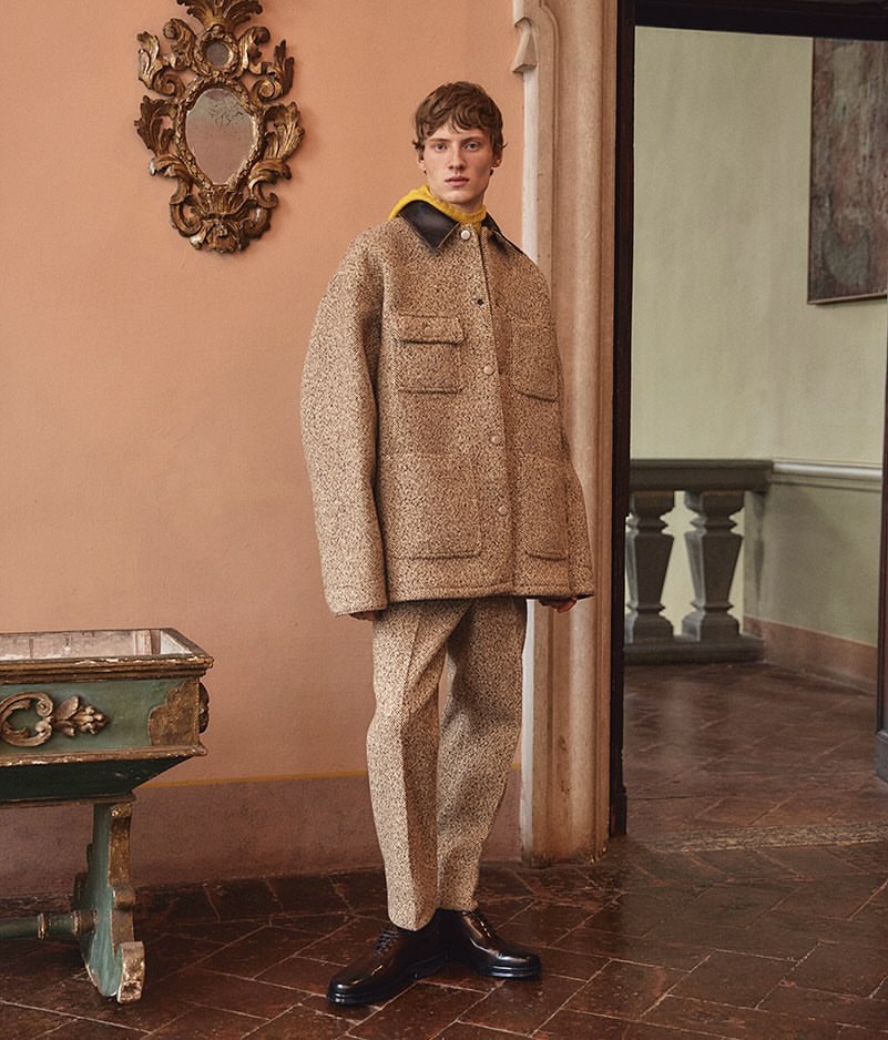 Fashionable man in a tweed jacket and pants poses indoors against a peach wall with an ornate mirror and a doorway in the background.
