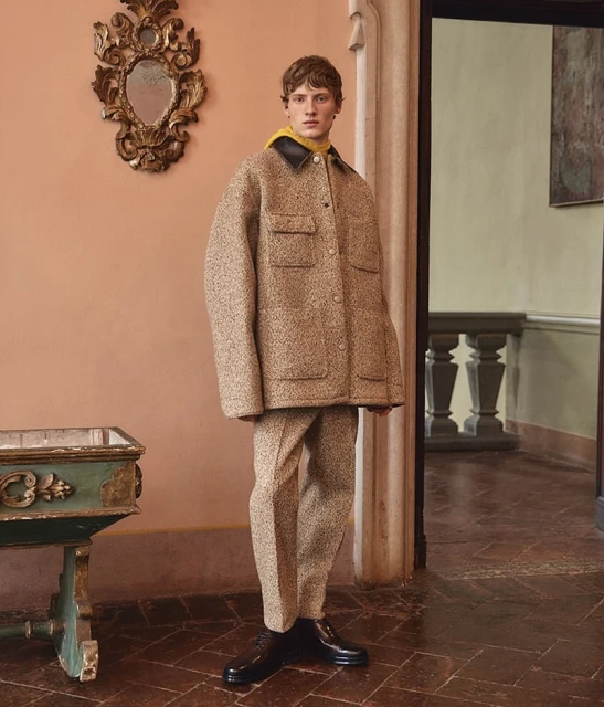 Fashionable man in a tweed jacket and pants poses indoors against a peach wall with an ornate mirror and a doorway in the background.