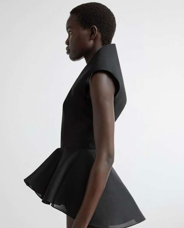 Side profile shot of a dark-skinned woman in a sculptural black top with dramatic flared peplum, against a bright white studio backdrop.