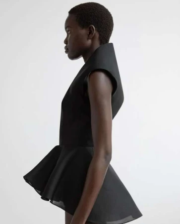 Side profile shot of a dark-skinned woman in a sculptural black top with dramatic flared peplum, against a bright white studio backdrop.