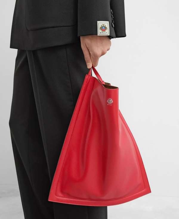 A person holds a red, triangular leather tote bag with the Bally logo, paired with a black suit in a studio setting.