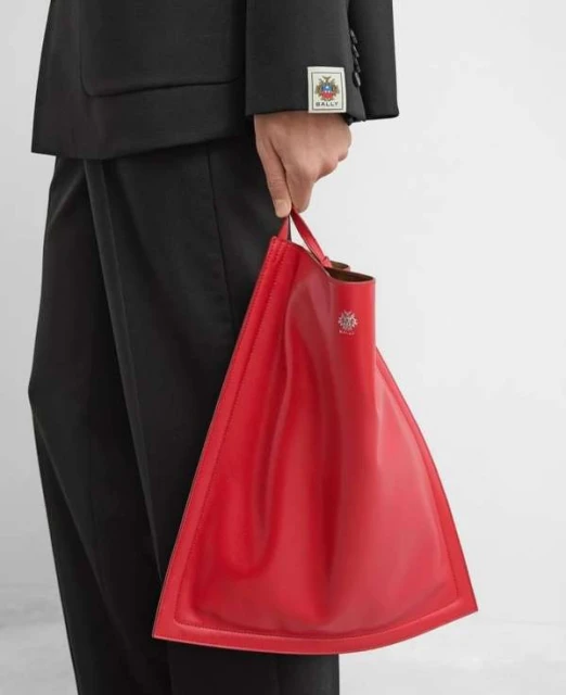 A person holds a red, triangular leather tote bag with the Bally logo, paired with a black suit in a studio setting.