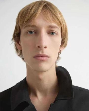 Portrait of a young man with blonde hair and blue eyes wearing a black jacket against a plain light background