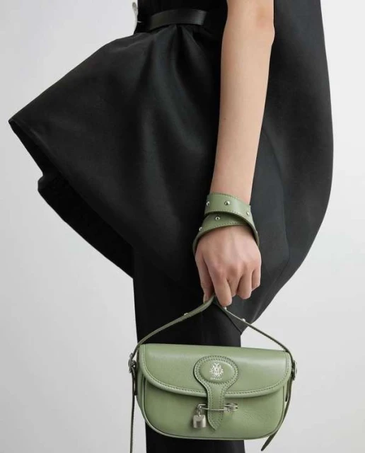 A fashionable woman in a black dress holds a green leather purse with metal hardware against a simple, neutral background.