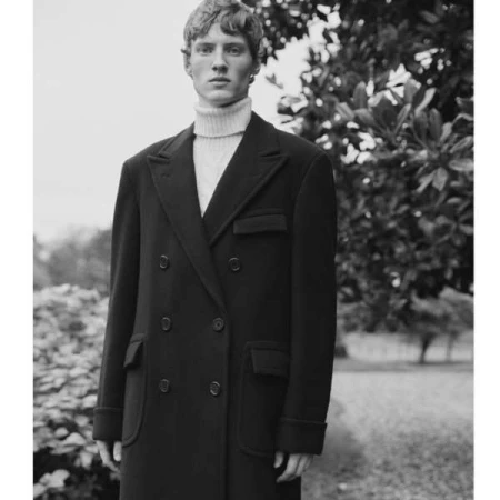 Monochrome portrait of a young man wearing a double breasted dark coat and a white turtleneck sweater outdoors.