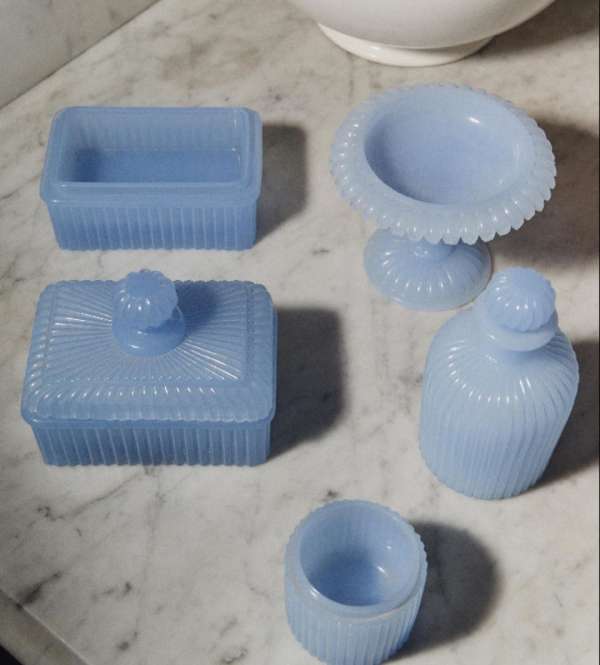 Assorted vintage light blue glass vanity set on marble surface featuring a soap dish, covered box, pedestal bowl, perfume bottle, and cup.