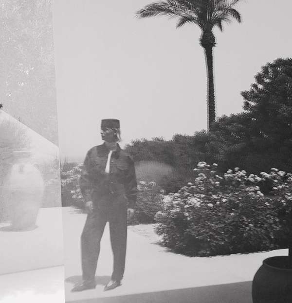 Monochrome photograph of a man in a uniform standing outdoors, with palm tree and greenery in the background, next to a decorative vase.