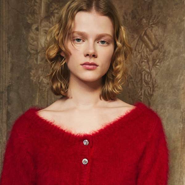Blonde woman poses wearing a red, off-the-shoulder mohair sweater with two buttons against a muted brown, textured background.