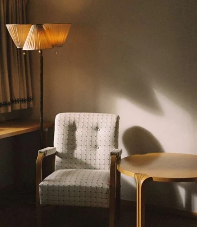 An armchair with patterned upholstery next to a wooden table and a standing lamp with three pleated shades in a room by Carlotta Manaigo