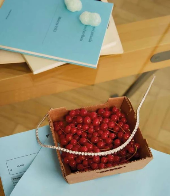 Close-up of a diamond tennis necklace draped over a box filled with vibrant red currants, placed on top of a light blue book.