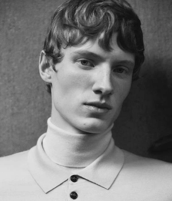 Black and white portrait of a fair-skinned adult male with wavy light brown hair, wearing a light-colored turtleneck shirt, indoors.