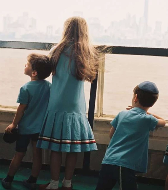 Three children in blue clothing stand near a railing with a distant urban skyline visible in the background, gazing out towards the water.