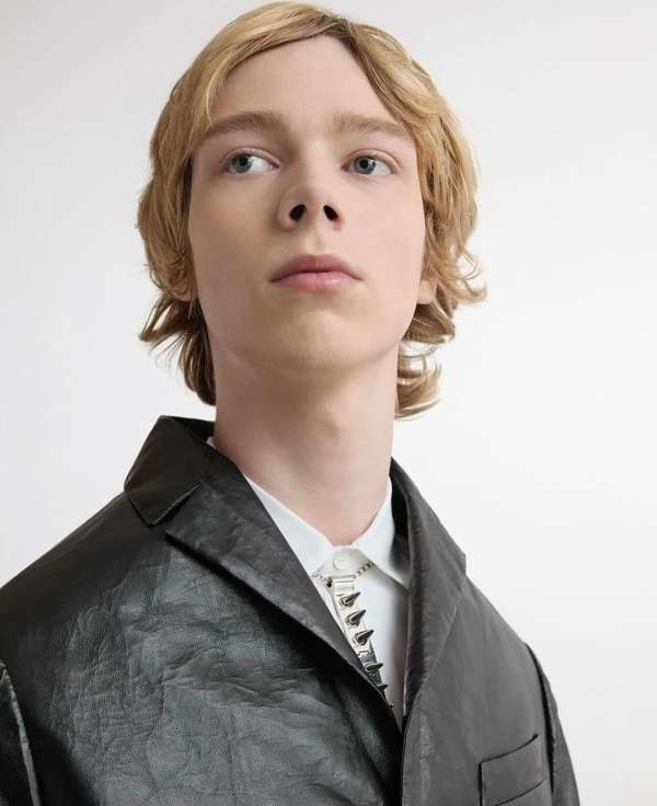 A young adult male model with blond hair poses in a black jacket and white shirt against a white backdrop in a fashion portrait.