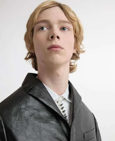 A young adult male model with blond hair poses in a black jacket and white shirt against a white backdrop in a fashion portrait.
