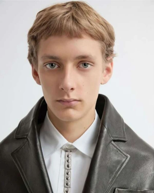 Close-up portrait of a young adult male with blonde hair wearing a white button-down shirt, a dark leather jacket, and a bolo tie.