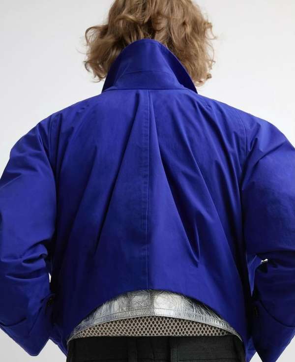 A person with blonde curly hair stands with their back to the camera, wearing a short blue jacket and a decorative silver belt.