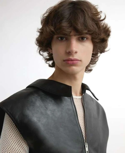Close-up studio portrait of a young adult male with brown curly hair wearing a black leather vest over a white mesh shirt. 