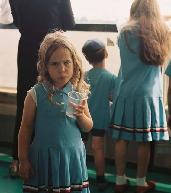A young girl with a serious expression is holding a plastic cup in front of her, surrounded by other children wearing blue outfits.