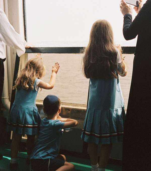 Three children look out a window at the water; two girls wear matching blue dresses with white and red trim, and one boy wears a blue shirt.