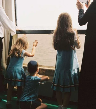 Three children look out a window at the water; two girls wear matching blue dresses with white and red trim, and one boy wears a blue shirt.