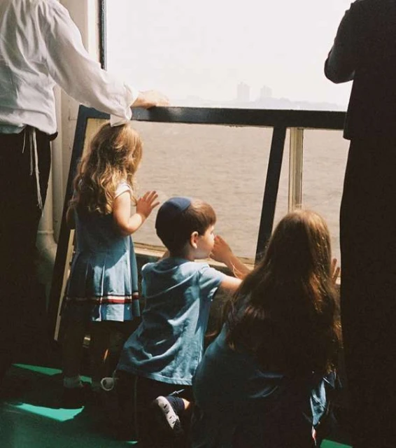 Three children and two adults gaze out a window on a boat, with a body of water and a distant cityscape visible through the glass.
