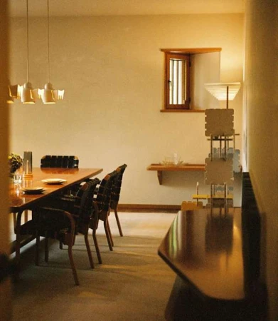 Interior shot of a dining room featuring a long wooden dining table, dark chairs, pendant lighting, and a small barred window.