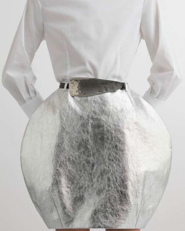 Fashionable woman from the back wearing a silver skirt, white blouse, and belt with a metallic buckle in a bright studio setting.