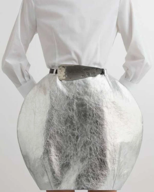 Fashionable woman from the back wearing a silver skirt, white blouse, and belt with a metallic buckle in a bright studio setting.