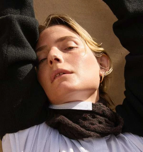 Close-up portrait of a light-skinned young adult with short blonde hair, wearing a striped shirt and dark sweater against a neutral backdrop.