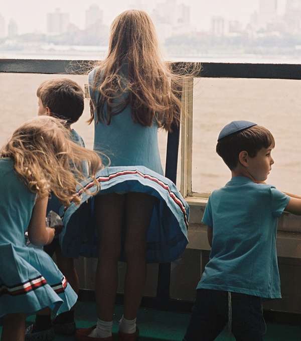 Five children, three girls and two boys, wearing blue dresses/shirts, standing on a boat looking out over the water and a city on the horizon.