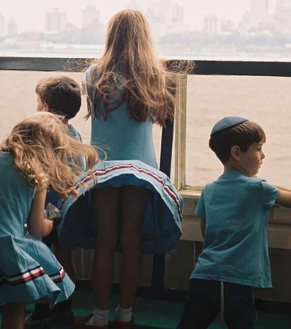 Five children, three girls and two boys, wearing blue dresses/shirts, standing on a boat looking out over the water and a city on the horizon.