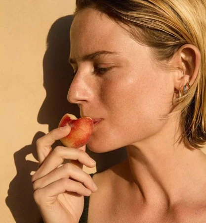 Close-up studio shot of an adult woman with short blonde hair, fair skin eating a peach against a warm beige background with shadows.