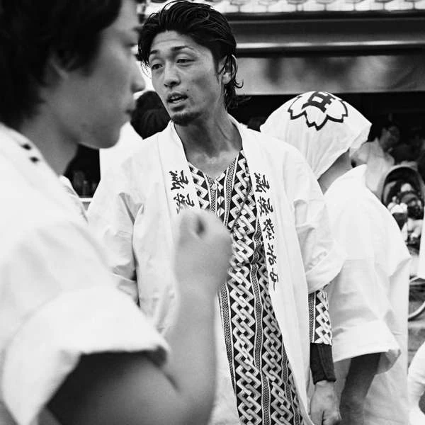 Monochrome portrait of two Asian men outdoors wearing traditional Japanese festival outfits with printed patterns, talking with each other.