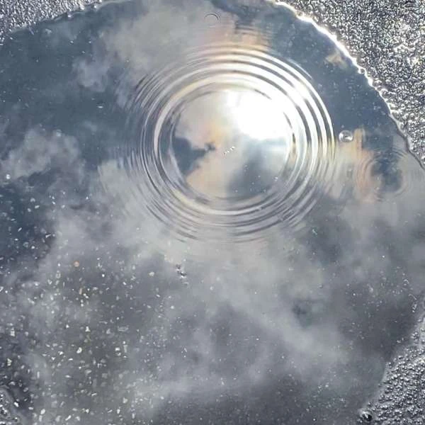 Ripples in a puddle reflecting a cloudy sky with a sun glare, creating a mesmerizing water surface texture.