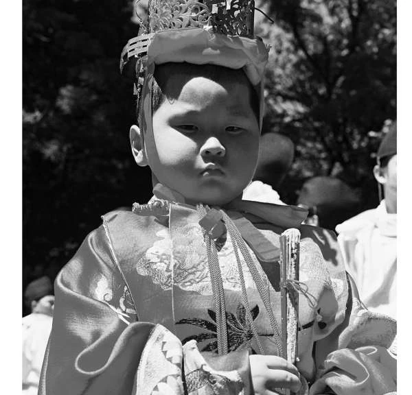 Monochrome close-up of a child wearing a decorative crown and costume, standing outdoors with trees in the background.