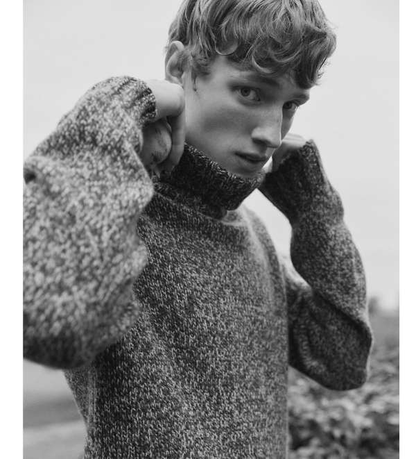 Monochrome portrait of a young adult male with curly blonde hair wearing a gray turtleneck sweater outdoors, posing for a fashion editorial.