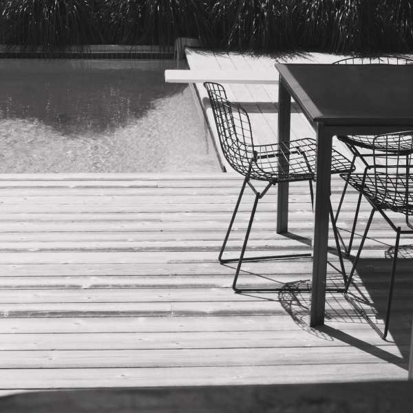 Monochrome image of an outdoor lounge area with chairs and a table on a wooden deck next to a swimming pool and garden.