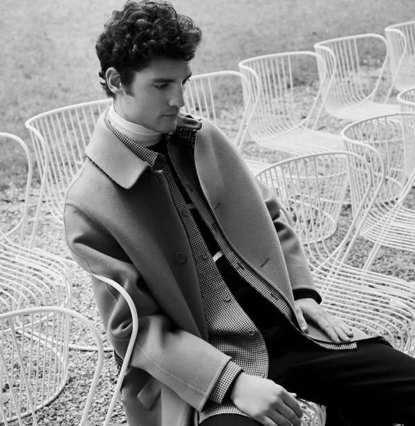 Black and white portrait of a man seated among many white chairs. The man wears a wool coat and turtleneck sweater, with a neutral expression.