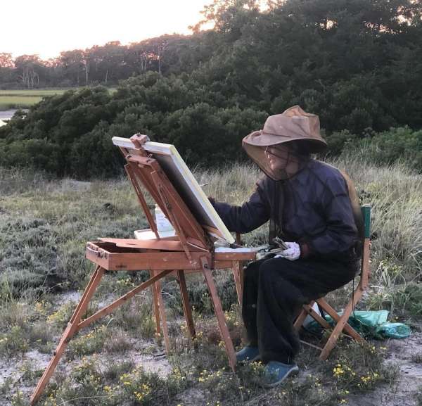 Adult painting en plein air in outdoor scene with artist's easel, brushes, canvas, protective hat with netting, and natural landscape.