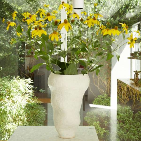 Close up of a white, textured vase holding a vibrant bouquet of yellow flowers with green leaves in an interior space.