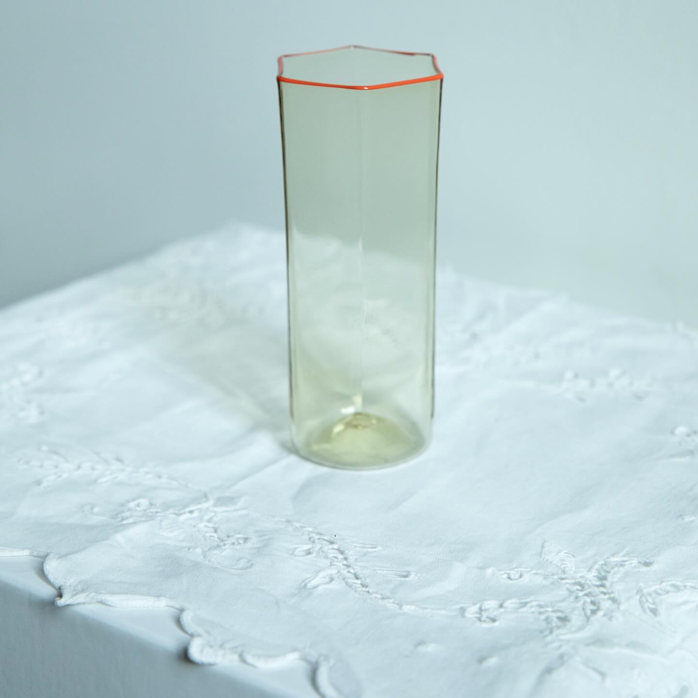 Tall, clear, hexagonal glass vase with an orange rim sits on a white embroidered tablecloth against a plain off-white background.