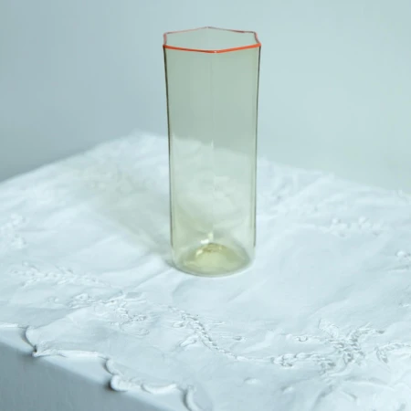 Tall, clear, hexagonal glass vase with an orange rim sits on a white embroidered tablecloth against a plain off-white background.