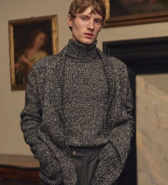 Adult male fashion model wearing a gray turtleneck sweater and dress pants posing indoors, with fine art paintings in the background.